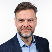 Auf dem Bild ist Julian Kamphausen zu erkennen. Er trägt einen dunkelblauen Anzug mit einem hellblauen Hemd. Außerdem hat er braun-graue, etwas länglichere Haare, die ihm zur Seite hängen. Zudem hat er blaue Augen und einen grau-braunen Bart.