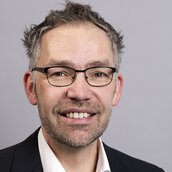 Auf dem Bild sieht man Manuel Fischer. Er hat kurze, grau-braune Haare und dunkle Augenbrauen. Außerdem trägt er eine schwarze Brille und lächelt mit Zähnen.