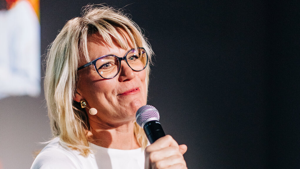 Auf dem Bild sieht man eine Frau mit schulterlangen, blonden Haaren. Sie trägt eine Brille und hält ein Mikrofon in der Hand.