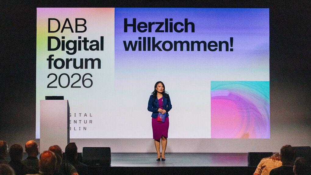 Auf dem Bild sieht man Thuy Hänelt-Do. Sie trägt ein lila farbendes Kleid mit einem dunkelblauen Blazer und schwarzen hohen Schuhen. Sie hat lange schwarze Haare und steht auf einer Bühne. Im Hintergrund sieht man eine Präsentationsfolie mit der Schrift "DAB Digitalforum 2026" und "Herzlich willkommen!"