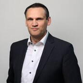 Auf dem Bild sieht man Dr. Norman Bernhardt. Er trägt einen schwarzen Anzug mit einem weißen Hemd. Außerdem hat er kurze, schwarze Haare und dunkle Augenbrauen.