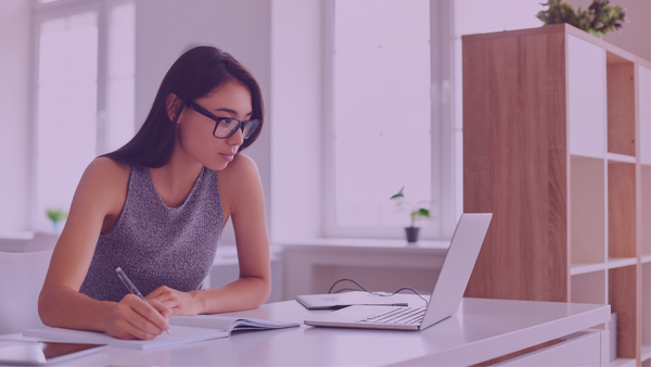 Auf dem Bild sieht man eine Frau, die an einem Schreibtisch sitzt und auf einen Laptop guckt. Vor ihr liegt ein Heft, in dem sie was reinschreibt. Sie hat lange dunkle Haare, trägt eine schwarze Brille und ein graues ärmelfreies Oberteilt. Das Bild ist zudem mit einem lila farbenden Flare überzogen.