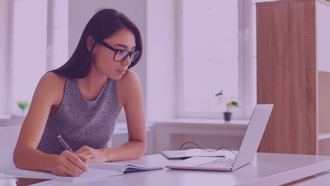 Auf dem Bild sieht man eine Frau, die an einem Schreibtisch sitzt und auf einen Laptop guckt. Vor ihr liegt ein Heft, in dem sie was reinschreibt. Sie hat lange dunkle Haare, trägt eine schwarze Brille und ein graues ärmelfreies Oberteilt. Das Bild ist zudem mit einem lila farbenden Flare überzogen.